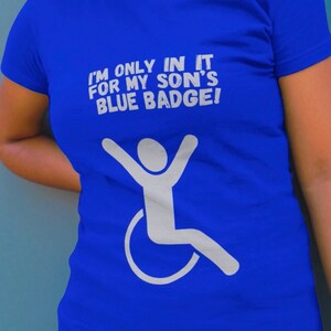 Peut inclure: Un t-shirt bleu royal avec du texte blanc qui dit "Je ne suis là que pour le badge bleu de mon fils !" et une silhouette blanche d'une personne en fauteuil roulant.