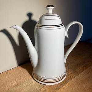 Op de afbeelding: Witte porseleinen koffiekan met een gouden rand rond de deksel en de basis. De kan heeft een rond lichaam en een tuit aan de ene kant.