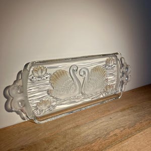 Puede incluir: Una bandeja de servir de vidrio transparente con un diseño en relieve de dos cisnes y nenúfares. La bandeja tiene dos asas a los lados.