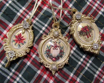 May include: Three ornate Christmas ornaments with gold frames and decorative embellishments. Each ornament features a different festive design: a poinsettia, a heart with berries, and a gift with a red bow. They hang from gold ribbons.