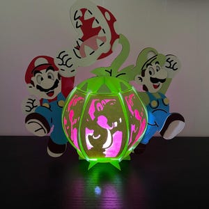 Puede incluir: Una l&aacute;mpara verde y blanca que se ilumina con forma de Planta Pira&ntilde;a de la serie de videojuegos Super Mario Bros. La l&aacute;mpara presenta dos figuras, una roja y una verde, de pie a cada lado de la planta. La l&aacute;mpara est&aacute; iluminada desde el interior, proyectando un brillo rosa.