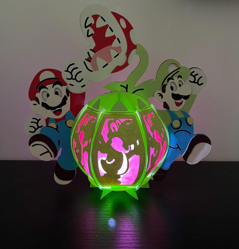 Puede incluir: Una l&aacute;mpara verde y blanca que se ilumina con forma de Planta Pira&ntilde;a de la serie de videojuegos Super Mario Bros. La l&aacute;mpara presenta dos figuras, una roja y una verde, de pie a cada lado de la planta. La l&aacute;mpara est&aacute; iluminada desde el interior, proyectando un brillo rosa.