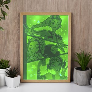 Peut inclure: Une impression d'art encadrée représentant une illustration éclairée en vert de trois personnages féminins stylisés. L'œuvre d'art est de style cartoon avec un cadre en bois clair. Le fond est vert clair avec des étoiles.