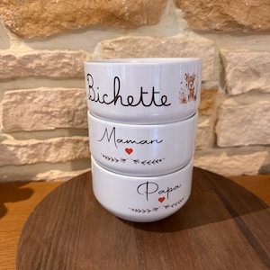 Peut inclure: Trois bols en céramique blanche empilés. Le bol supérieur porte l'inscription "Bichette", le bol du milieu "Maman" avec un cœur rouge, et le bol inférieur "Papa". Les bols sont sur un plateau en bois.