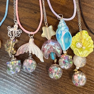Könnte beinhalten: Eine Sammlung von sechs Halsketten mit verschiedenen Anhängern, darunter ein mit Steinsplittern gefülltes Herz, ein Oktopus, eine Meerjungfrauenschwanzflosse, ein Stern, eine Mondsichel, eine Muschel und eine gelbe Muschel, ergänzt durch kleine Glaskugeln.
