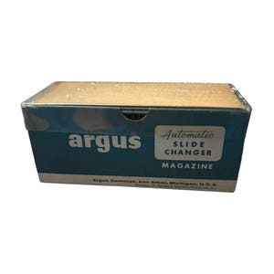 Puede incluir: Una caja de revista de cambiador de diapositivas automático Argus vintage. La caja es de color verde azulado y bronceado con texto blanco que dice "argus", "Automatic Slide Changer" y "Magazine". La caja también incluye el texto "Argus Cameras, Ann Arbor, Michigan, U.S.A."