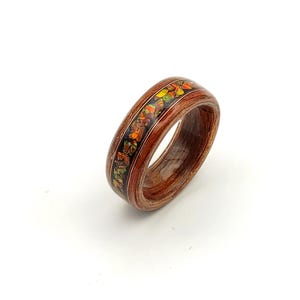 Può includere: Anello in legno fatto a mano con un'incastonatura vibrante di pietre preziose arancioni, gialle e verdi. L'anello presenta una fascia in legno marrone scuro e una base intarsiata nera, creando un contrasto sorprendente. Un gioiello unico.