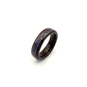 Op de afbeelding: Een houten ring met een donkerbruine band en een centrale inleg van blauw en goudkleurig materiaal. De ring staat tegen een witte achtergrond en toont zijn gepolijste afwerking en ingewikkelde ontwerp. Een uniek sieraad.