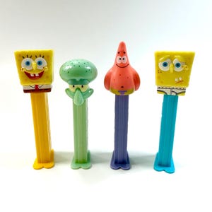 May include: Four plastic Pez candy dispensers featuring characters from SpongeBob SquarePants. The set includes two yellow SpongeBob figures, one light green Squidward figure, and one pink Patrick Star figure, each on a matching colored stem.