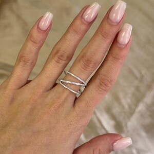Puede incluir: Un anillo plateado con un dise&ntilde;o entrecruzado, adornado con peque&ntilde;as piedras brillantes. El anillo se lleva en una mano con u&ntilde;as cuidadas. El dise&ntilde;o del anillo presenta m&uacute;ltiples bandas que se cruzan.