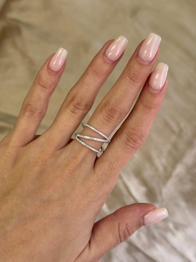 Puede incluir: Un anillo plateado con un dise&ntilde;o entrecruzado, adornado con peque&ntilde;as piedras brillantes. El anillo se lleva en una mano con u&ntilde;as cuidadas. El dise&ntilde;o del anillo presenta m&uacute;ltiples bandas que se cruzan.