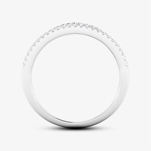 Puede incluir: Un anillo de metal blanco con una banda curva, engastado con peque&ntilde;as piedras preciosas claras en la parte superior. El anillo se muestra sobre un fondo liso y claro, resaltando su superficie pulida y el brillo de las piedras. Una joya.