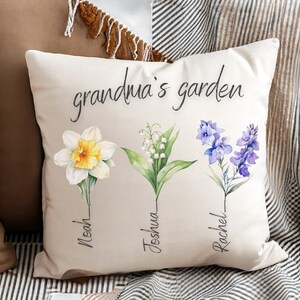 Puede incluir: Coj&iacute;n cuadrado beige con el texto "grandma's garden" en escritura negra. El coj&iacute;n presenta ilustraciones en acuarela de tres flores: un narciso, lirios de los valles y delfinios azules, cada una con un nombre debajo.