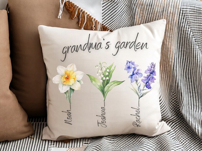 Puede incluir: Coj&iacute;n cuadrado beige con el texto "grandma's garden" en escritura negra. El coj&iacute;n presenta ilustraciones en acuarela de tres flores: un narciso, lirios de los valles y delfinios azules, cada una con un nombre debajo.