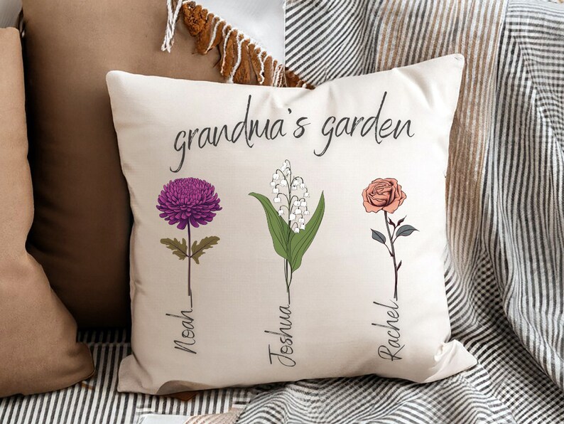 May include: White throw pillow with the text "grandma's garden" and three floral illustrations. The flowers are a purple chrysanthemum, a cluster of white lily of the valley, and a pink rose. Each flower has a name written below it: Noah, Joshua, and Rachel.