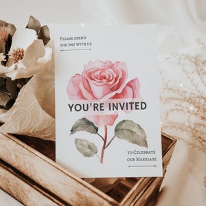 Puede incluir: Una tarjeta de invitaci&oacute;n de boda con una ilustraci&oacute;n de rosa rosa en acuarela. La tarjeta dice "YOU'RE INVITED", "PLEASE SPEND THE DAY WITH US" y "TO CELEBRATE OUR MARRIAGE". La tarjeta est&aacute; en una caja de madera con arreglos florales.