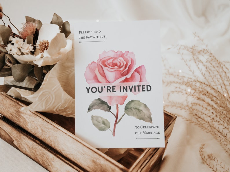 Puede incluir: Una tarjeta de invitaci&oacute;n de boda con una ilustraci&oacute;n de rosa rosa en acuarela. La tarjeta dice "YOU'RE INVITED", "PLEASE SPEND THE DAY WITH US" y "TO CELEBRATE OUR MARRIAGE". La tarjeta est&aacute; en una caja de madera con arreglos florales.