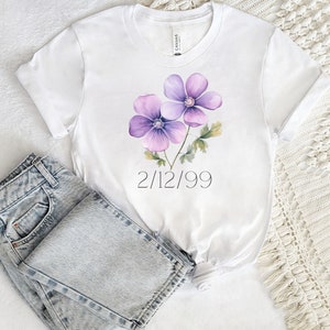 Puede incluir: Camiseta blanca con un dise&ntilde;o de acuarela de dos flores moradas y hojas verdes. La fecha "2/12/99" est&aacute; impresa debajo. Vaqueros de mezclilla de lavado claro doblados junto a la camiseta.