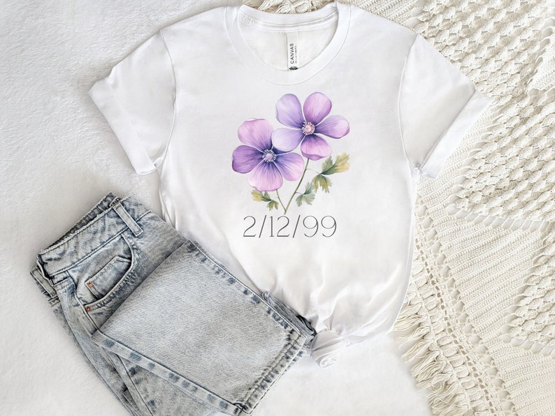 Puede incluir: Camiseta blanca con un dise&ntilde;o de acuarela de dos flores moradas y hojas verdes. La fecha "2/12/99" est&aacute; impresa debajo. Vaqueros de mezclilla de lavado claro doblados junto a la camiseta.