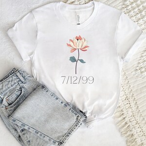 May include: White t-shirt with a floral design and the date 7/12/99 printed on the front. The shirt is folded and laid on a white surface with a pair of light blue jeans.