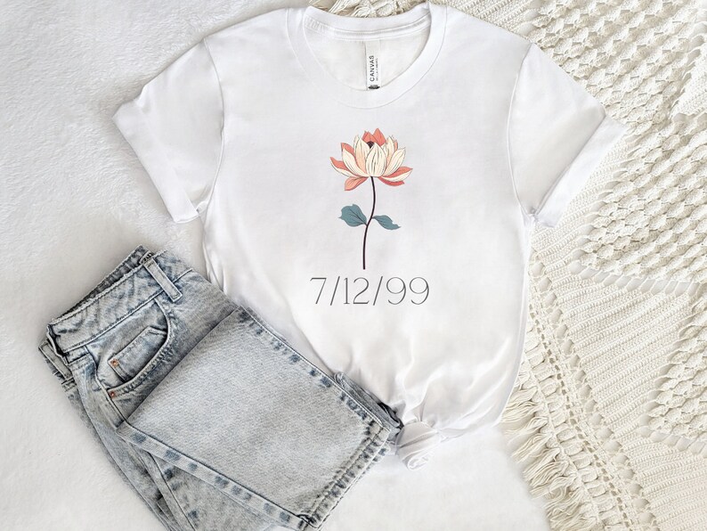 May include: White t-shirt with a floral design and the date 7/12/99 printed on the front. The shirt is folded and laid on a white surface with a pair of light blue jeans.
