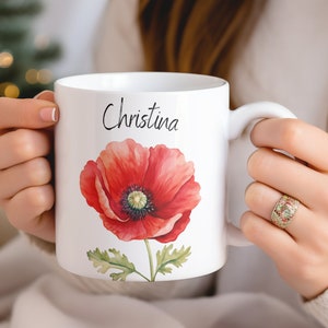 Puede incluir: Taza de cer&aacute;mica blanca con una ilustraci&oacute;n de amapola roja vibrante con hojas verdes y el nombre "Christina" en escritura negra. La taza tiene un asa blanca y es sostenida por una persona.