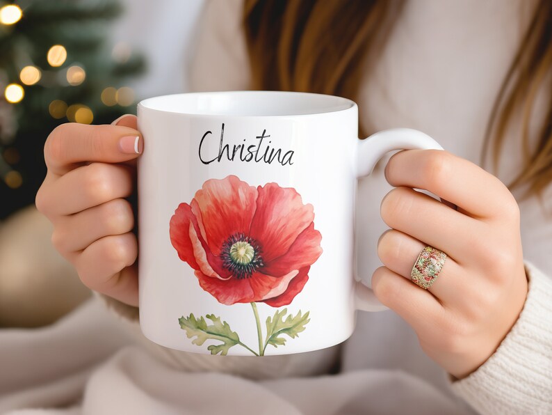 Puede incluir: Taza de cer&aacute;mica blanca con una ilustraci&oacute;n de amapola roja vibrante con hojas verdes y el nombre "Christina" en escritura negra. La taza tiene un asa blanca y es sostenida por una persona.