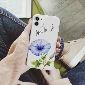 Puede incluir: Una funda de tel&eacute;fono blanca con una ilustraci&oacute;n de acuarela de una flor de gloria de la ma&ntilde;ana azul con hojas verdes y el texto "Libra for Life" en escritura negra. La funda est&aacute; en las manos de una persona.