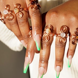 May include: A set of six copper wire rings with various semi-precious stones. The rings are all in a similar style, with a twisted wire design and a single stone in the center. The stones are a mix of brown, white, and clear.