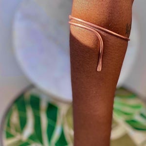 May include: A copper arm cuff with a minimalist design. The cuff wraps around the upper arm and features a curved, teardrop-shaped accent. The jewelry is worn on a brown arm, with a blurred background.