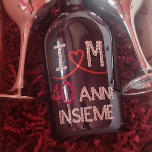 Puede incluir: Una botella negra con una cinta roja y las palabras "I ♥ M 40 ANNI INSIEME" decoradas con pedrería. Dos copas de champán plateadas con las palabras "G.  L.O.B.O. J & M" están a cada lado de la botella. La botella y las copas están en una caja con papel picado rojo.