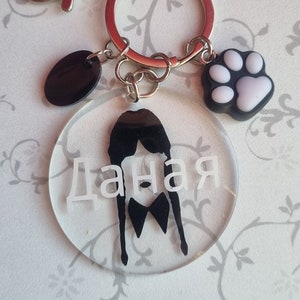 May include: A clear round keyring with a black silhouette of a person with the name "Даная" written in white. The keyring has a silver metal ring and a silver metal clasp. There is a black round charm and a white paw print charm attached to the ring.