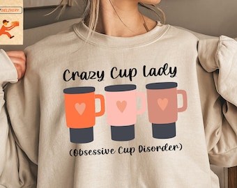 May include: A beige sweatshirt with the text "Crazy Cup Lady (Obsessive Cup Disorder)" and a graphic of three colorful mugs with hearts.