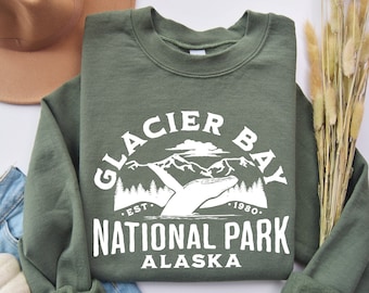 May include: A green sweatshirt with a white graphic design that says "Glacier Bay National Park Alaska" with a mountain range and a hand reaching out to the mountains. The text also says "Est. 1980."