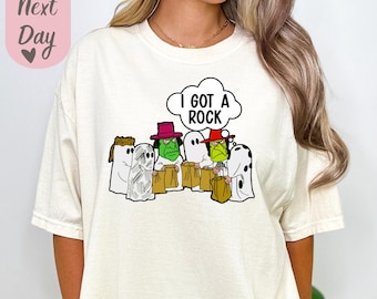 May include: A white t-shirt with a graphic of ghosts holding bags of candy. The text "I got a rock" is in a thought bubble above the ghosts.
