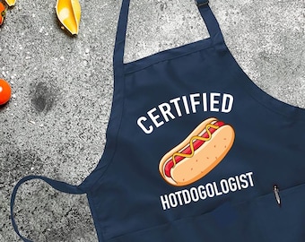 May include: A navy blue apron with a white pocket and the text "CERTIFIED HOTDOGOOLOGIST" with a cartoon hot dog graphic.