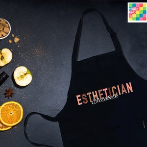 Pode incluir: Avental preto com o texto "ESTHETICIAN Elizabeth" em letras douradas rosadas. O avental &eacute; cercado por v&aacute;rios ingredientes, incluindo ma&ccedil;&atilde;s, laranjas, paus de canela e anis estrelado.
