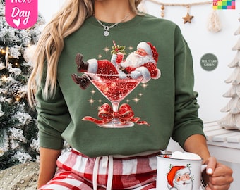 May include: A military green sweatshirt features a graphic of Santa Claus relaxing in a red cocktail glass. The sweatshirt is paired with red and white plaid pajama pants. A white mug with a Santa Claus face is held in the hand.
