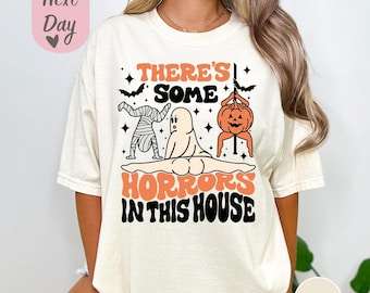 May include: A white t-shirt with a black and orange graphic that says "There's some horrors in this house". The graphic features a mummy, a ghost, and a pumpkin.