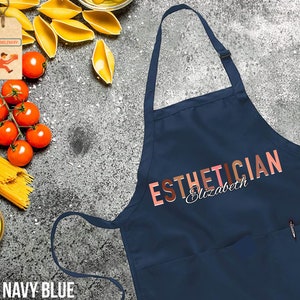 Pode incluir: Avental azul marinho com um bolso grande e o texto "ESTHETICIAN Elizabeth" em letras coloridas.