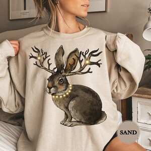 Op de afbeelding: Een beige sweatshirt met een grafische print van een bruin konijn met gewei en een string lichtjes om zijn nek. De tekst "SAND" is gedrukt op de rechteronderhoek van het sweatshirt.