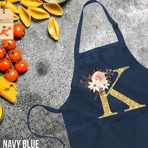 May include: Navy blue kitchen apron with a large gold glitter letter K and floral design. The letter K is surrounded by white and pink flowers.
