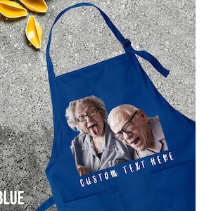 May include: Royal blue apron with a custom photo print of an elderly couple making silly faces. The apron has two pockets and the text "CUSTOM TEXT HERE" in white. Pasta and tomatoes are in the background.