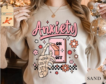 May include: A beige sweatshirt with the text "Anxiety" in pink lettering. A cartoon skeleton hand is turning a pink switch labeled "ON" and "OFF". The switch is surrounded by pink flowers and stars.
