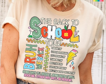 May include: A cream-colored t-shirt featuring a colorful graphic with the text THE BACK TO SCHOOL TOUR, BRUH WE BACK, and a list of tasks including LAMINATIN, LESSON PLANNING, and CAFFEINE DRINKIN, plus a red apple and yellow lightning bolt.