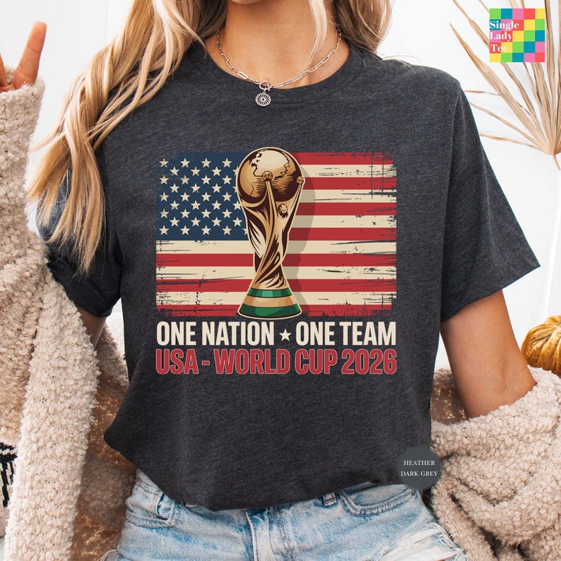 A person wearing a black t-shirt with a graphic design featuring the FIFA World Cup trophy and the text "One Nation, One Team USA-World Cup 2026" printed on it.