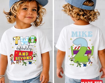 May include: A white short-sleeve t-shirt featuring a graphic print with the text "TWO infinity AND BEYOND" and "MIKE" above a large number "2". The design includes colorful cartoon characters and stars, with a blue baseball cap worn by the wearer.