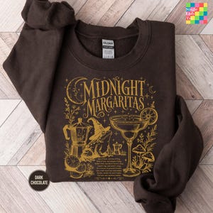 May include: A dark chocolate brown crewneck sweatshirt featuring a gold graphic print. The design includes the text "MIDNIGHT MARGARITAS," a crescent moon, a frog wearing a witch hat, a margarita glass, mushrooms, and a moka pot coffee maker.