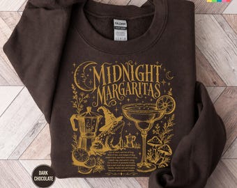 May include: A dark chocolate brown crewneck sweatshirt featuring a gold graphic print. The design includes the text "MIDNIGHT MARGARITAS," a crescent moon, a frog wearing a witch hat, a margarita glass, mushrooms, and a moka pot coffee maker.
