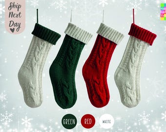 May include: Four knitted Christmas stockings hanging from a string. The stockings are white with a green, red, and white cuff. The stockings are decorated with a cable knit pattern.